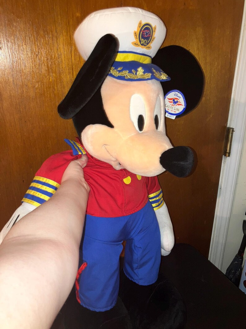 May include: A large plush Mickey Mouse doll wearing a white captain's hat with gold accents and a blue and gold band. Mickey is dressed in a red jacket with gold buttons and blue trousers. A Disney Cruise Line tag is attached.