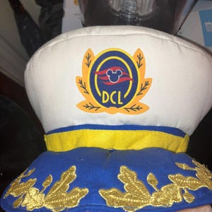 May include: A white captain's hat with a blue and yellow brim. The hat features a gold emblem with the text "DCL" and a Mickey Mouse silhouette. The hat is likely a souvenir or accessory.