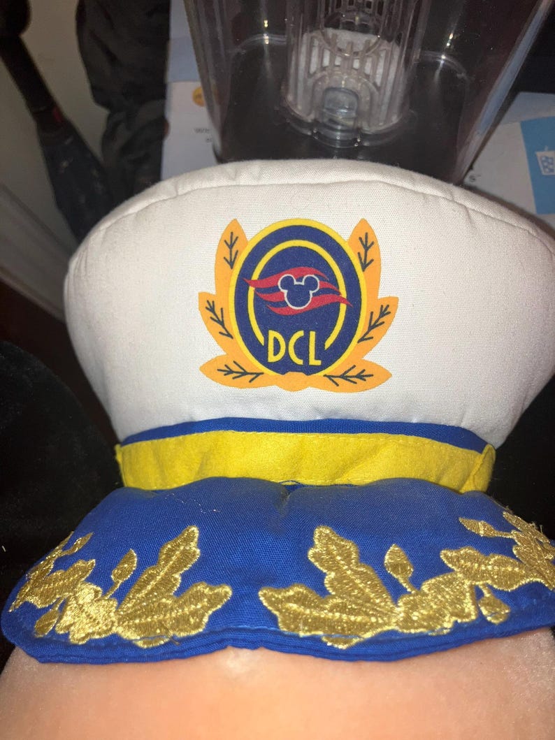 May include: A white captain's hat with a blue and yellow brim. The hat features a gold emblem with the text "DCL" and a Mickey Mouse silhouette. The hat is likely a souvenir or accessory.