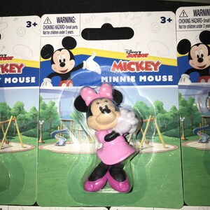 Peut inclure: Une figurine en plastique de Minnie Mouse de la s&eacute;rie Disney Junior Mickey Mouse. Minnie porte une robe rose et des gants blancs. Elle tient une fleur blanche. La figurine est emball&eacute;e sur une carte avec un fond vert et une sc&egrave;ne de terrain de jeu. La carte porte une &eacute;tiquette d'avertissement qui dit "RISQUE D'&Eacute;TOUFFEMENT - Petites pi&egrave;ces. Ne convient pas aux enfants de moins de 3 ans."