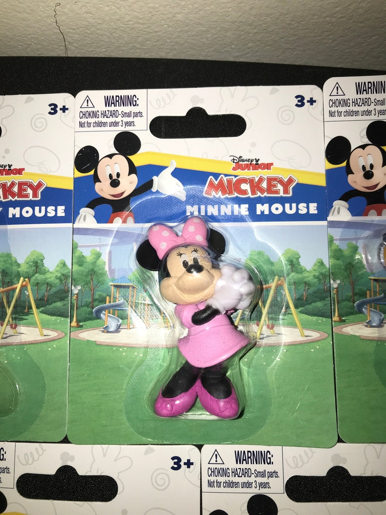 Peut inclure: Une figurine en plastique de Minnie Mouse de la s&eacute;rie Disney Junior Mickey Mouse. Minnie porte une robe rose et des gants blancs. Elle tient une fleur blanche. La figurine est emball&eacute;e sur une carte avec un fond vert et une sc&egrave;ne de terrain de jeu. La carte porte une &eacute;tiquette d'avertissement qui dit "RISQUE D'&Eacute;TOUFFEMENT - Petites pi&egrave;ces. Ne convient pas aux enfants de moins de 3 ans."