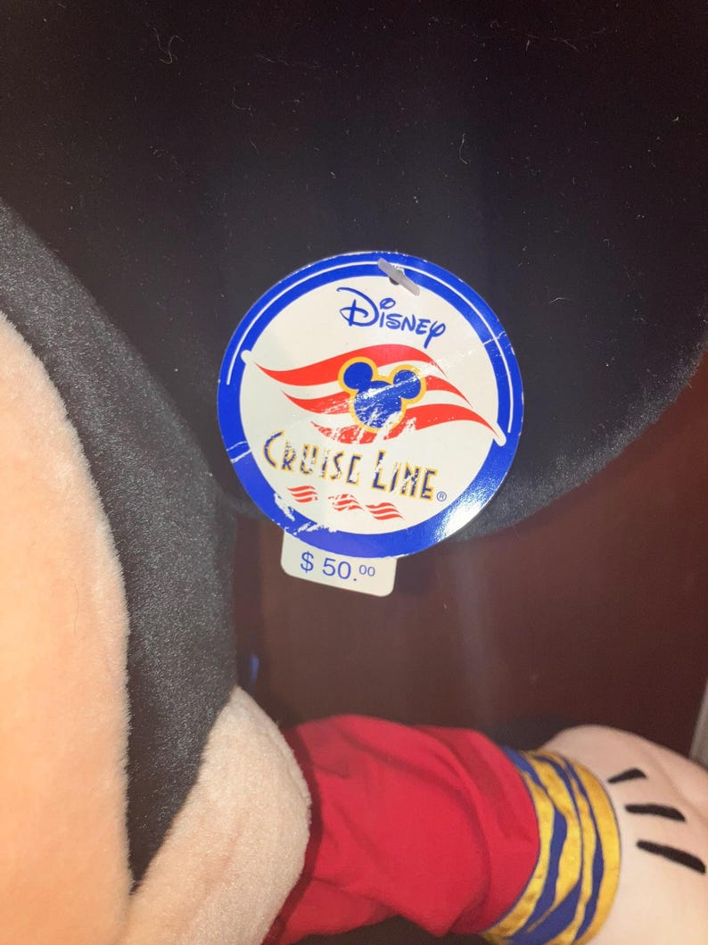 May include: A Mickey Mouse soft toy in a red shirt with yellow and blue details. The toy has a black head and ears, and a Disney Cruise Line tag attached.