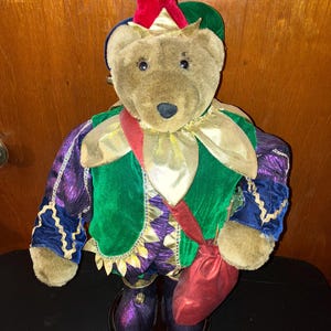 May include: A plush teddy bear dressed in a colorful jester costume. The bear wears a red and green hat, a green and purple velvet outfit with gold accents, and carries a red pouch. The bear is standing on a dark, round base.