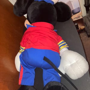 May include: A large plush toy of a cartoon mouse, viewed from the back. The mouse wears a red jacket with gold stripes, blue trousers, a white sailor hat with a blue band, and black shoes. The mouse's ears are black, and its hands are white.