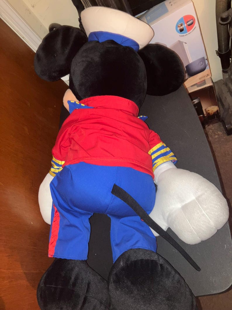 May include: A large plush toy of a cartoon mouse, viewed from the back. The mouse wears a red jacket with gold stripes, blue trousers, a white sailor hat with a blue band, and black shoes. The mouse's ears are black, and its hands are white.