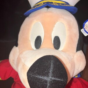 May include: A plush Mickey Mouse doll wearing a white captain's hat with a blue brim and gold detailing. The doll has a red shirt and a large black nose. The hat has the letters "DCL" on it.