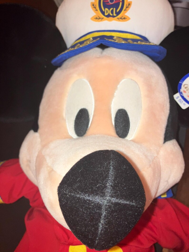 May include: A plush Mickey Mouse doll wearing a white captain's hat with a blue brim and gold detailing. The doll has a red shirt and a large black nose. The hat has the letters "DCL" on it.