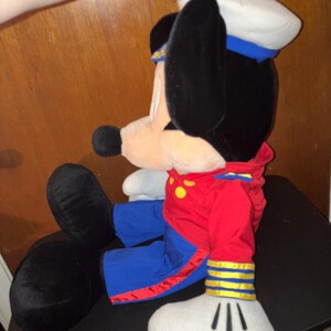 May include: A plush Mickey Mouse doll in a sailor outfit. The doll has black ears, a white sailor hat with a blue band, and a red jacket with yellow buttons. The doll's trousers are blue, and its gloves are white.