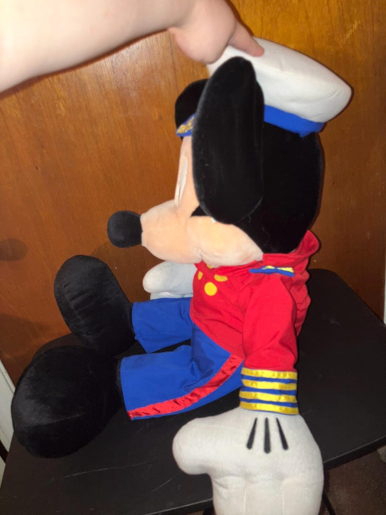 May include: A plush Mickey Mouse doll in a sailor outfit. The doll has black ears, a white sailor hat with a blue band, and a red jacket with yellow buttons. The doll's trousers are blue, and its gloves are white.