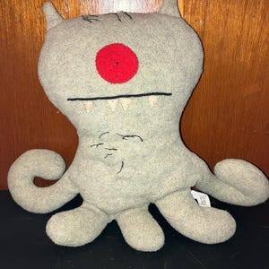 May include: A gray plush toy with a large red eye, small horns, and multiple tentacle-like appendages. The toy has a stitched mouth with teeth and stitched eyebrows. The toy is on a black surface.