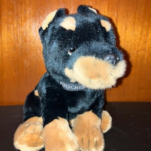 May include: A plush toy dog, primarily black with tan markings on the paws, ears, and snout. The dog has a black nose and a collar with the word "Pinscher" on it. The toy is sitting upright.