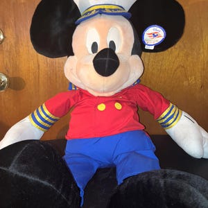 May include: A Mickey Mouse soft toy dressed in a captain's uniform. The toy features a white hat with a gold emblem, a red shirt with yellow buttons, blue trousers, and black ears and feet.