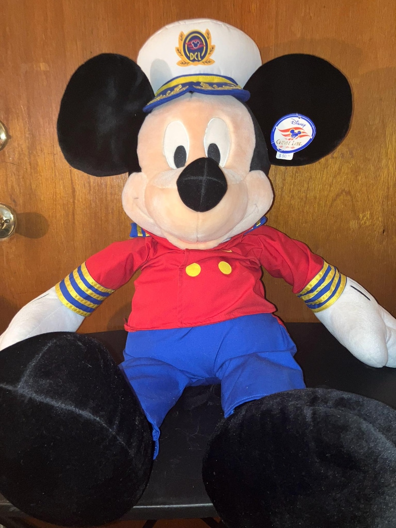 May include: A Mickey Mouse soft toy dressed in a captain's uniform. The toy features a white hat with a gold emblem, a red shirt with yellow buttons, blue trousers, and black ears and feet.