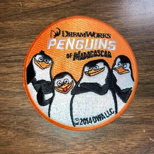 May include: Round orange patch featuring the DreamWorks Penguins of Madagascar. The patch shows four cartoon penguins in black, white, and orange. Text includes "DreamWorks Penguins of Madagascar" and "©2014 DWA LLC".