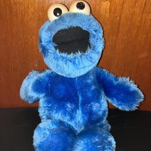 May include: A plush Cookie Monster toy, featuring a vibrant blue fur body, large white eyes with black pupils, and a wide open black mouth. The toy is seated, with its arms and legs visible, and is made of soft, fuzzy material.