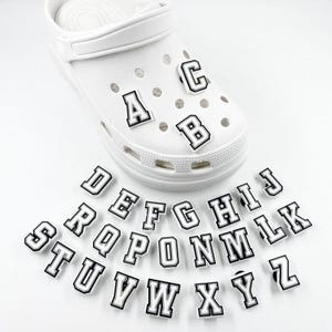 May include: White clog shoe with black and white letter charms. The letters A, B, and C are attached to the shoe. Additional letters are scattered around the shoe. The letters have a black outline.