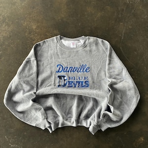 Pode incluir: Um moletom cinza mesclado com a inscrição bordada em azul "Danville Blue Devils". O moletom tem gola redonda e mangas compridas. O texto está em letra cursiva.