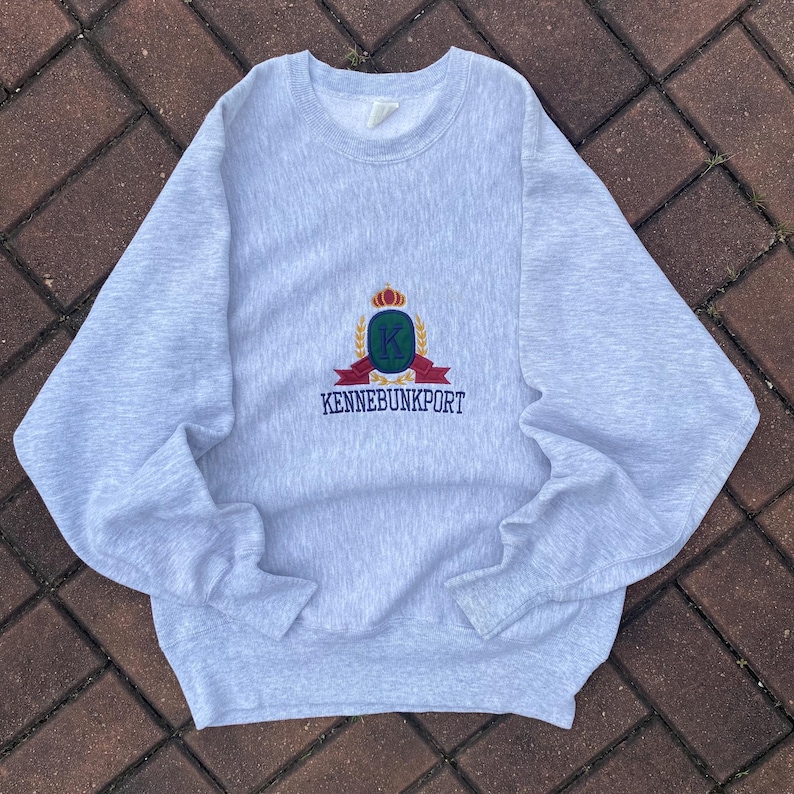 May include: A gray sweatshirt with a crest embroidered on the front. The crest features a crown, a letter K, and the text "KENNEBUNKPORT".