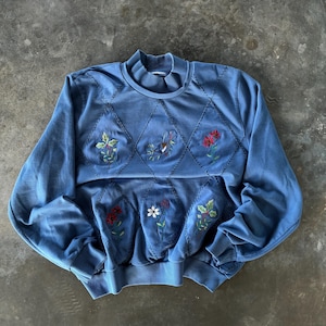 Puede incluir: Una sudadera azul con diseños florales bordados. La sudadera presenta un cuello alto y mangas largas. La parte delantera de la sudadera tiene un patrón de diamantes con flores y hojas bordadas.