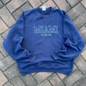 Puede incluir: Una sudadera azul marino con el texto "MIAMI FLORIDA" en letras blancas con contorno.