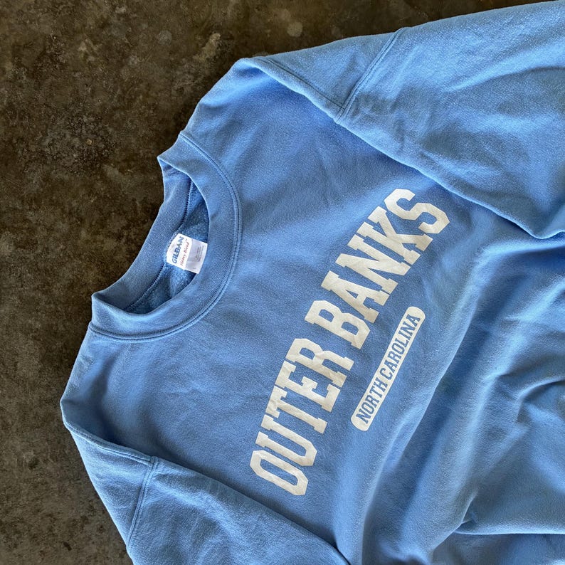 以下が含まれることがあります： 大きな白い文字で「OUTER BANKS」と書かれた、水色のスウェットシャツ。「NORTH CAROLINA」の文字が下に小さく書かれています。柔らかい素材のスウェットシャツです。