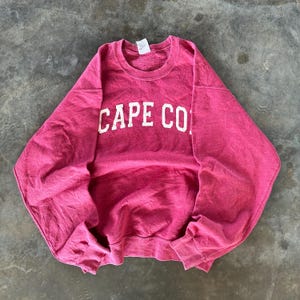 May include: A crimson-colored sweatshirt with the text "CAPE CO" in white block letters. The crew neck sweatshirt is made of a soft material and has long sleeves. The garment is laid flat on a concrete surface.