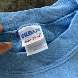 以下が含まれることがあります： ライトブルーのGildan Heavy Blendスウェットシャツ。「50%コットン、50%ポリエステル、ホンジュラス製」と記載されたタグとサイズ「L」が含まれています。スウェットシャツは無地のライトブルーです。