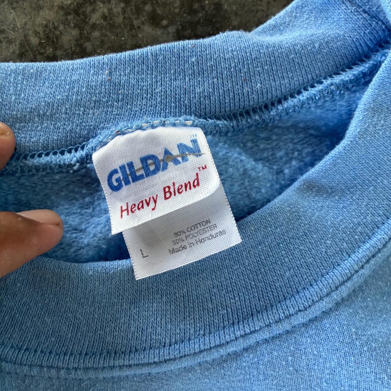 以下が含まれることがあります： ライトブルーのGildan Heavy Blendスウェットシャツ。「50%コットン、50%ポリエステル、ホンジュラス製」と記載されたタグとサイズ「L」が含まれています。スウェットシャツは無地のライトブルーです。