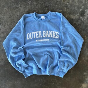 以下が含まれることがあります： 胸に白い文字で「OUTER BANKS」と書かれた、水色のクルーネックのスウェットシャツ。その下に「NORTH CAROLINA」の文字が小さく表示されています。スウェットシャツは柔らかい素材で作られています。