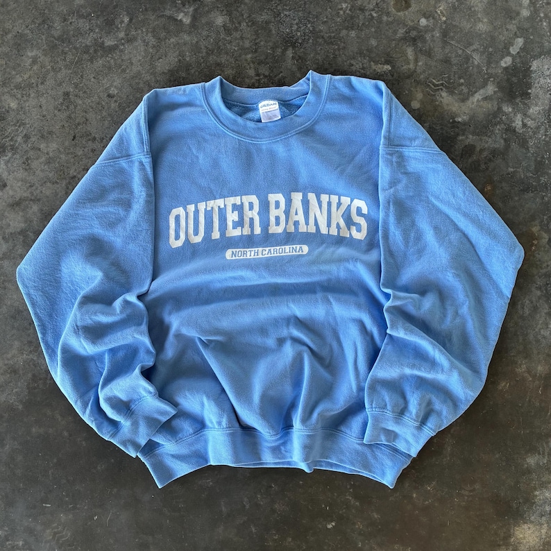 以下が含まれることがあります： 胸に白い文字で「OUTER BANKS」と書かれた、水色のクルーネックのスウェットシャツ。その下に「NORTH CAROLINA」の文字が小さく表示されています。スウェットシャツは柔らかい素材で作られています。