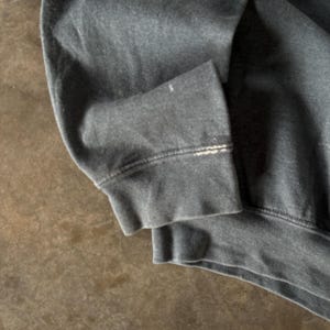 May include: A dark charcoal gray long-sleeve sweatshirt featuring a ribbed cuff with a visible horizontal line of wear and slight fraying along the seam. The fabric appears to be a soft cotton blend with a faded, worn-in texture throughout the sleeve.