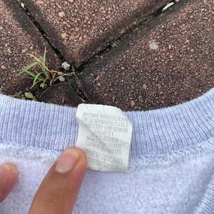 May include: A white sweatshirt with a light blue trim around the neck. The care label is attached to the inside of the neck and reads "Machine wash delicate cycle separately-cold, tumble dry low remove promptly, Do not bleach, Do not iron or dry clean if decorated, Garment designed shrink to size."