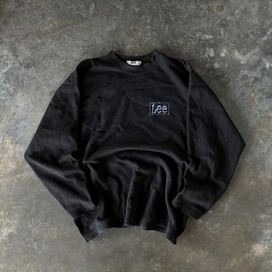 May include: Black long-sleeve sweatshirt with a crew neck. The sweatshirt features a small, embroidered "Lee" logo on the chest. The fabric appears to be a soft, worn material. The overall style is casual and vintage.
