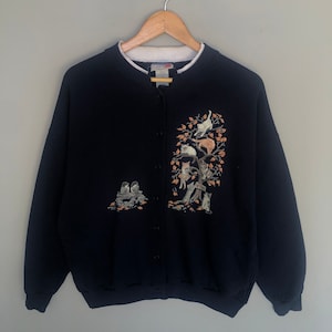 Puede incluir: Una chaqueta de punto azul marino con botones y cuello blanco. La chaqueta presenta un diseño bordado de gatos trepando a un árbol con pájaros en una rama debajo.