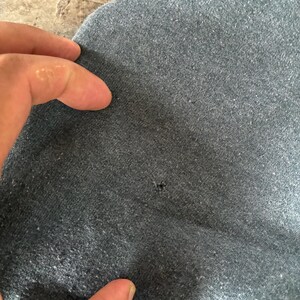 May include: A dark gray heathered fabric garment featuring a small, singular hole in the center of the material. The textile has a textured, slightly worn appearance with visible pilling across the surface, indicating a soft, knit construction.