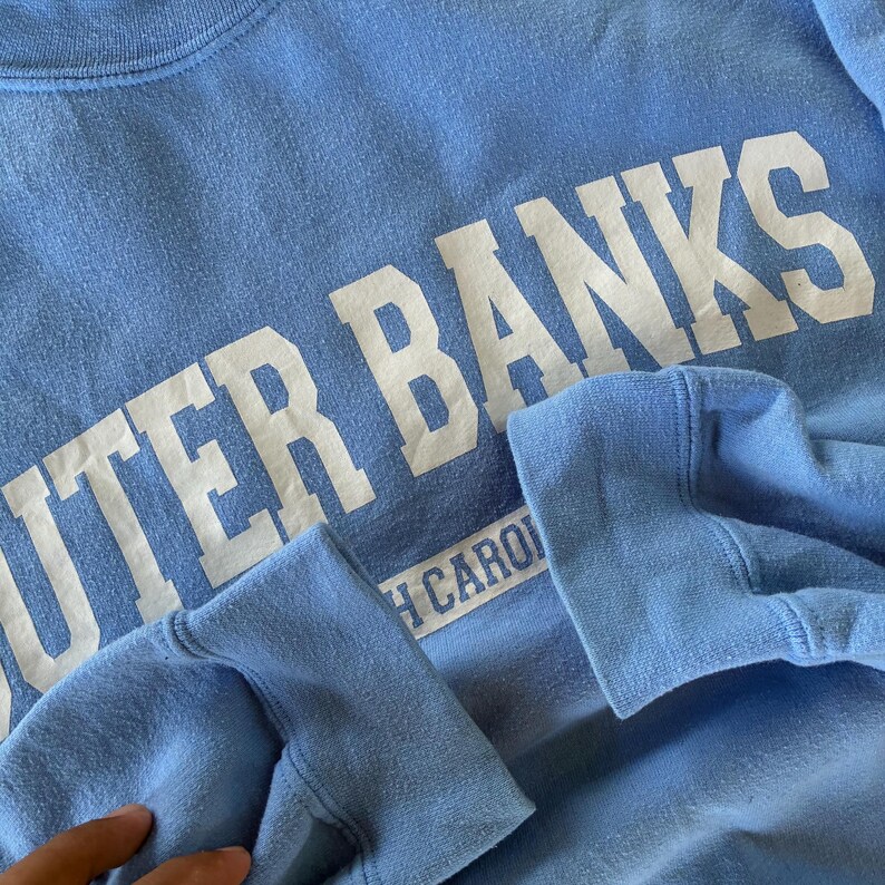 以下が含まれることがあります： 大きな白い文字で「OUTER BANKS」と書かれた水色のスウェットシャツ。テキストの下には「NORTH CARO」の文字が見えます。袖口は折り返されています。