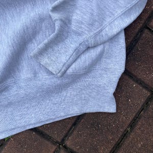 May include: A close-up of the gray knit fabric of a sweater. The fabric is soft and textured, with a ribbed knit pattern. The sweater has a long sleeve and a ribbed cuff.