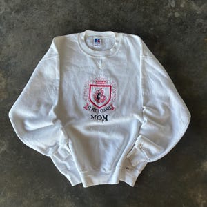 May include: White crew neck sweatshirt with a red and pink crest design. The crest features the words "St. Peter Chanel" and the word "MOM" embroidered in red. The sweatshirt is a Russell brand.