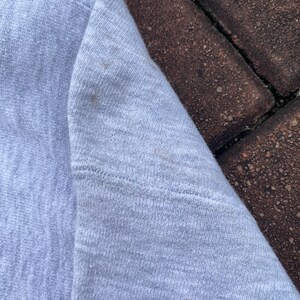 May include: Close-up of a light gray sweatshirt fabric. The fabric has a subtle texture and a slightly faded appearance.