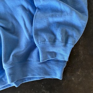 以下が含まれることがあります： 水色のスウェットシャツの袖のクローズアップ。生地は柔らかく、わずかに摩耗しているように見え、テクスチャが見えます。袖口はリブ編みで、ステッチが見えます。全体的な色は、無地の淡い青色です。