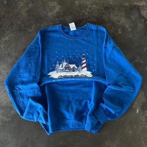 May include: A blue sweatshirt featuring a winter scene with a lighthouse and a house covered in snow. The lighthouse has red and white stripes. White snowflakes are falling from the sky. The sweatshirt is made of a soft material.