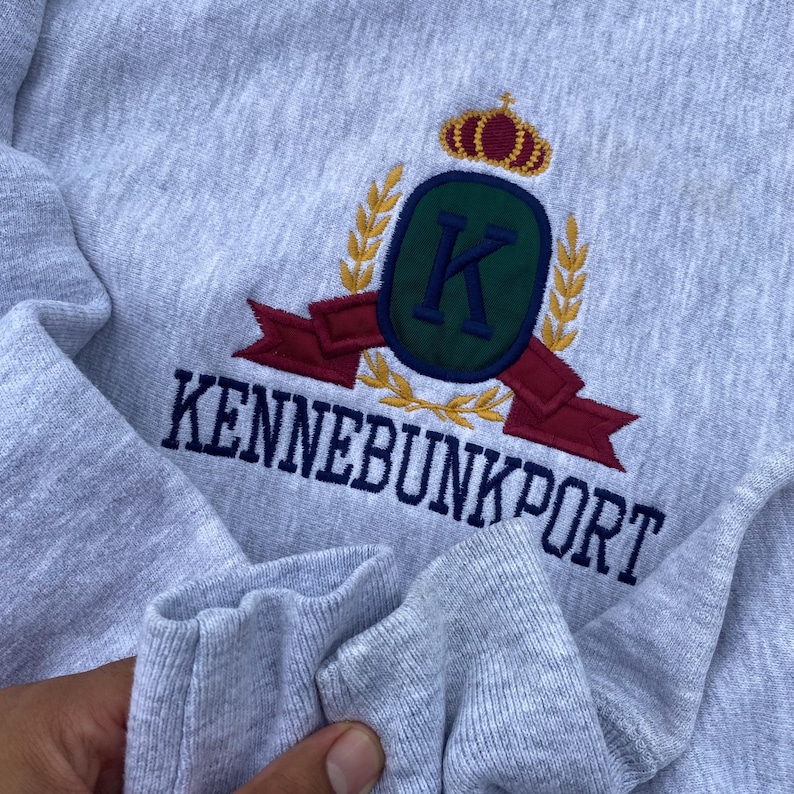 May include: Gray sweatshirt with a navy blue embroidered logo featuring a crown, the letter K, and the text "Kennebunkport".