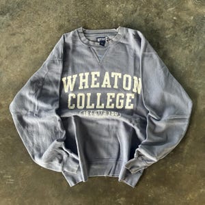 May include: A faded blue sweatshirt featuring the text "WHEATON COLLEGE" in white lettering. Below the text is the number "1860". The sweatshirt has a crew neck and long sleeves.