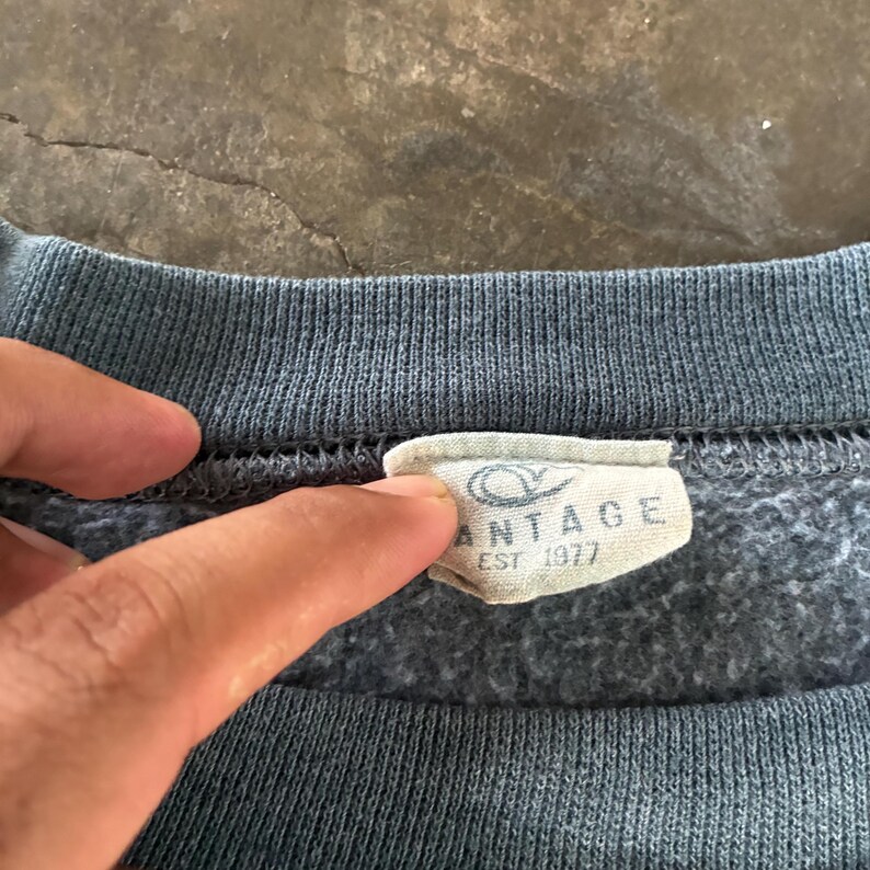 May include: A dark gray crewneck sweatshirt featuring a ribbed collar and a white fabric interior tag. The tag displays a stylized logo above the text VANTAGE and EST 1977 in black lettering. The garment has a soft, fleeced texture on the inside.