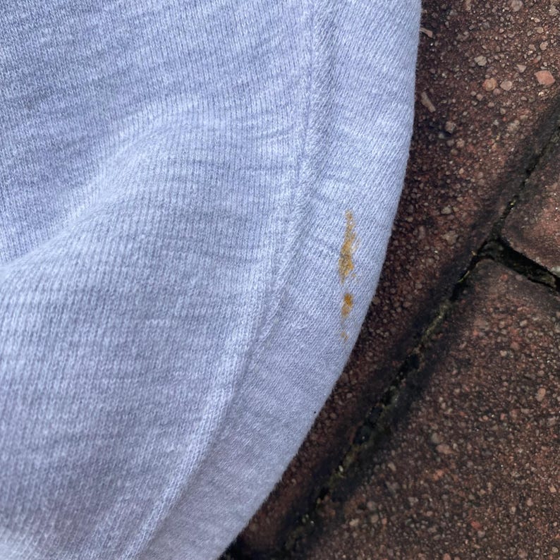 May include: A close-up of a light gray sweatshirt with a small brown stain on the fabric.