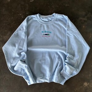 Op de afbeelding: Een lichtblauwe sweatshirt met de tekst "MT RAINIER NATIONAL PARK" geborduurd in groen, blauw en rood. De top met lange mouwen en ronde hals is gemaakt van een zachte stof.