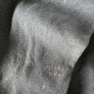 May include: A dark charcoal gray fabric with a visible knit texture and a soft, slightly fuzzy surface. The material displays subtle variations in tone and light wear, featuring small, light-colored fibers scattered across the dark surface.