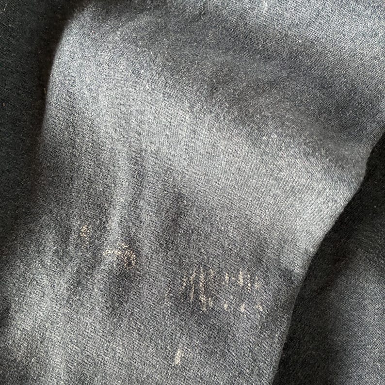 May include: A dark charcoal gray fabric with a visible knit texture and a soft, slightly fuzzy surface. The material displays subtle variations in tone and light wear, featuring small, light-colored fibers scattered across the dark surface.