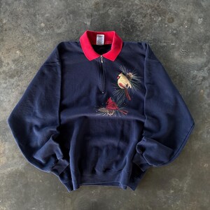 May include: Navy blue sweatshirt with a red collar and a quarter-zip closure. The sweatshirt features an embroidered design of two cardinal birds perched on pine branches. The birds are red and tan, with detailed feathering.