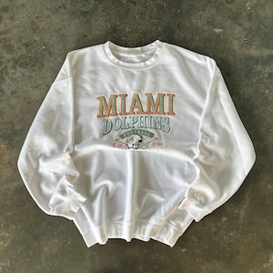 Puede incluir: Sudadera blanca de cuello redondo con el texto "MIAMI DOLPHINS" en letras turquesas y naranjas. El diseño incluye un gráfico de casco de fútbol y el texto "FOOTBALL Est. 1966". La sudadera está hecha de un material suave y cómodo.